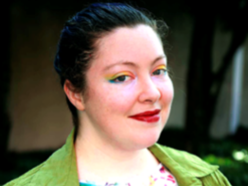 A smiling fat white person with long dark hair pulled back into a braid, wearing multicolor eye makeup, dark red lipstick, and a shirt with abstract spatters of bright colors.