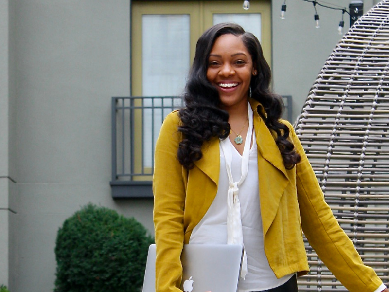 A young black woman smiling while walking towards the camera holding a laptop, wearing a white shirt and yellow blazer.